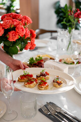 Hand holding plate with assorted bruschettas on festive table with red roses, glassware, and...