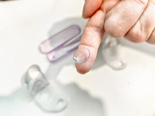 Fototapeta premium Clear contact lens being held on fingertip with storage cases in the background