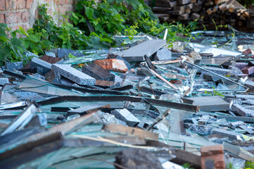Broken glass pieces lie amidst debris and overgrown plants in a forsaken alleyway, showing signs of...