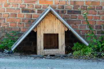 A rustic wooden dog house sits against a red brick wall, partially hidden by lush green plants in a...
