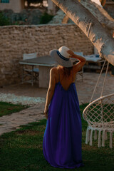 Woman in Blue Dress Walking Towards Outdoor Retreat

A woman in a flowing blue dress and striped hat walks across green grass towards a white building and macrame swing, bathed in warm su