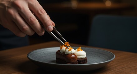 Chef is using tweezers to decorate a dessert with orange flowers on a dark plate on a wooden table
