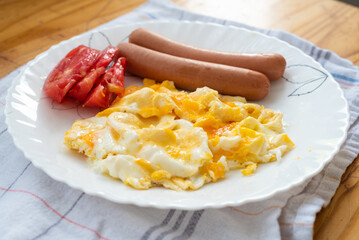 Traditional breakfast plate with scrambled eggs, sausages, fresh tomato slices, and bread, served on a white plate with a homey kitchen background.
