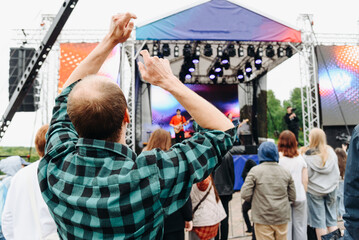 music festival in city park, crowd of people backview, rock group performing on stage, green trees and summer day