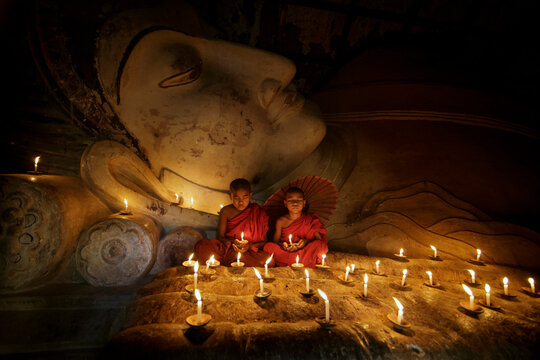 Two Buddhist monks dressed in traditional robes (kasaya) sitting in front of Buddha statue surrounded by candles praying, Myanmar