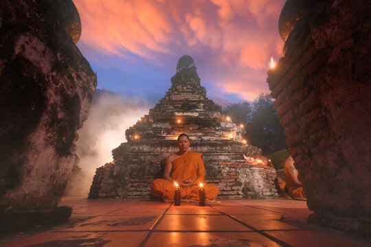 Buddhist monk in traditional orange robe (kasaya) sitting on the floor of a temple meditating, Thailand