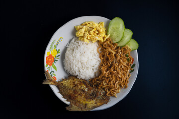 Flat lay of a plate with rice, fried fish, fried noodles, sliced omelette, and cucumber on a dark background