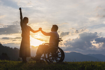 Disabled handicapped young man in wheelchair raised hands with his care helper in sunset.Silhouette