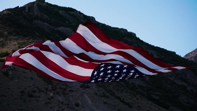 Cinematic drone shot lowering below flowing American flag in desert canyon at dusk - super slow motion