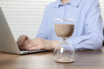 Deadline. Woman with laptop working at wooden table in office, focus on hourglass