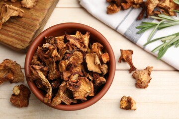 Dried chanterelle mushrooms in bowl and rosemary on light wooden table, flat lay