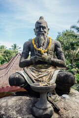 Stone statue of a Balinese man in a meditative pose, adorned with a yellow flower garland....