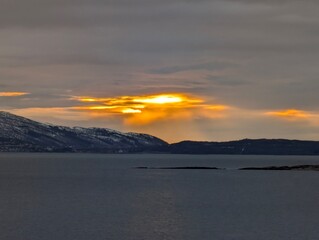Sun breaking through thick clouds over distant snowy hills and sea