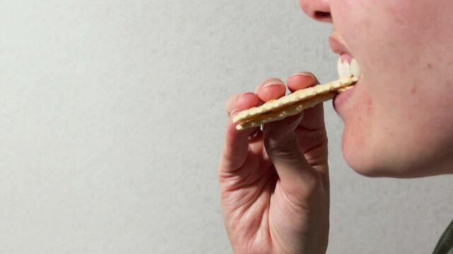 Side view of a woman taking a bit of a sandwich made with two saltine crackers captured against a neutral gray background