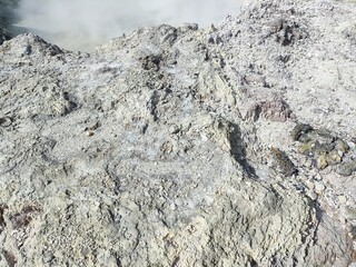 An active fumarole or crater area, featuring light-colored sulfurous earth and vapor rising into the air under a vast blue sky.