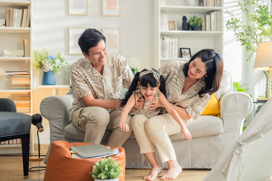 Happy Asian family enjoying quality time in their modern living room. Cheerful young parents playfully carry and bond with their daughter while laughing together on the sofa, creating joyful moments a