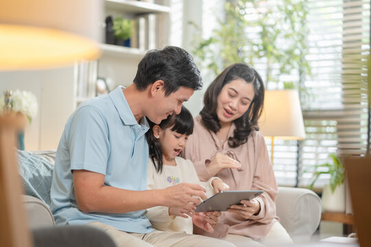 Happy Asian family with father mother and daughter using digital tablet laptop for home-based learning education while watching movies cartoons sitting together on living room sofa creating joyful - Powered by Adobe