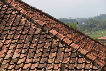 Fototapeta premium Close-up of aged red-brown rustic roof tiles with moss and a blurred village backdrop in soft daylight. Architecture texture concept