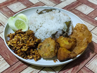 Complete Indonesian uduk rice served with tempe orek, egg, fried chicken, tofu fritters and cucumber slices on a white plate with a brown wooden patterned tablecloth in the background.