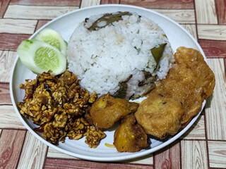 Complete Indonesian uduk rice served with tempe orek, egg, fried chicken, tofu fritters and cucumber slices on a white plate with a brown wooden patterned tablecloth in the background.