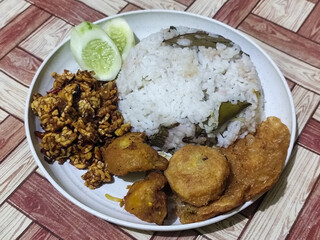 Complete Indonesian uduk rice served with tempe orek, egg, fried chicken, tofu fritters and cucumber slices on a white plate with a brown wooden patterned tablecloth in the background.