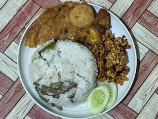 Complete Indonesian uduk rice served with tempe orek, egg, fried chicken, tofu fritters and cucumber slices on a white plate with a brown wooden patterned tablecloth in the background.