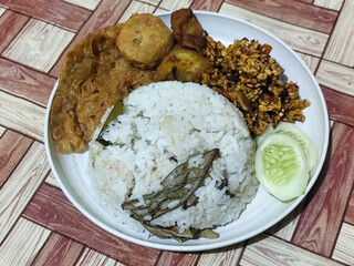 Complete Indonesian uduk rice served with tempe orek, egg, fried chicken, tofu fritters and cucumber slices on a white plate with a brown wooden patterned tablecloth in the background.