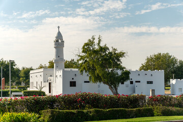 Traditional white mosque with a minaret stands in contrast to modern skyscrapers of West Bay in Doha, Qatar. Green park and blue sky create a peaceful urban landscape.