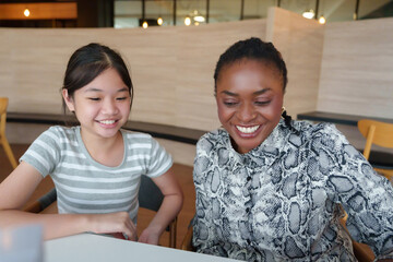 Smiling Diverse teacher and Asian girl student discussing lessons and studying together happily in...