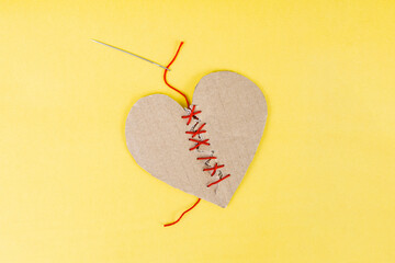 Broken cardboard heart sewed together with a needle and red thread on yellow background, marriage counseling concept