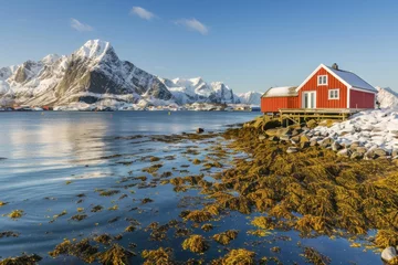 Papier peint photo Villes / Voyage Stunning red cabin by the snowy shoreline with majestic mountains in the background during a cloudy day in winter  © Olena