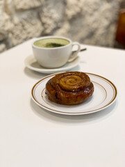 Delicious glazed cinnamon roll served on a white plate, paired with a cup of frothy matcha latte. Cozy café vibes with a rustic stone wall background.