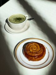 Golden cinnamon roll with glossy glaze served on a plate beside a creamy matcha latte. Bright sunlight casts artistic shadows on the white table.