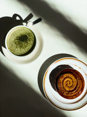 Golden cinnamon roll with glossy glaze served on a plate beside a creamy matcha latte. Bright sunlight casts artistic shadows on the white table.