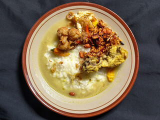 Bubur ayam. A warm Indonesian dish of chicken porridge with savory yellow sauce, chicken pieces, peanuts and orek tempeh served completely on a brown plate.