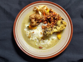 Bubur ayam. A warm Indonesian dish of chicken porridge with savory yellow sauce, chicken pieces, peanuts and orek tempeh served completely on a brown plate.
