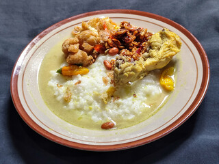 Bubur ayam. A warm Indonesian dish of chicken porridge with savory yellow sauce, chicken pieces, peanuts and orek tempeh served completely on a brown plate.