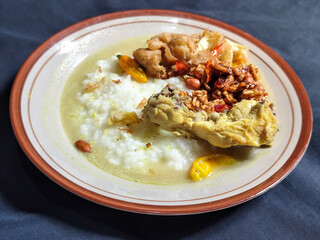 Bubur ayam. A warm Indonesian dish of chicken porridge with savory yellow sauce, chicken pieces, peanuts and orek tempeh served completely on a brown plate.
