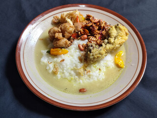 Bubur ayam. A warm Indonesian dish of chicken porridge with savory yellow sauce, chicken pieces, peanuts and orek tempeh served completely on a brown plate.