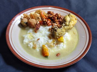 Bubur ayam. A warm Indonesian dish of chicken porridge with savory yellow sauce, chicken pieces, peanuts and orek tempeh served completely on a brown plate.