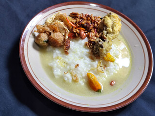 Bubur ayam. A warm Indonesian dish of chicken porridge with savory yellow sauce, chicken pieces, peanuts and orek tempeh served completely on a brown plate.