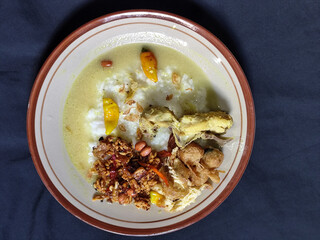 Bubur ayam. A warm Indonesian dish of chicken porridge with savory yellow sauce, chicken pieces, peanuts and orek tempeh served completely on a brown plate.