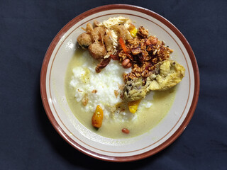 Bubur ayam. A warm Indonesian dish of chicken porridge with savory yellow sauce, chicken pieces, peanuts and orek tempeh served completely on a brown plate.