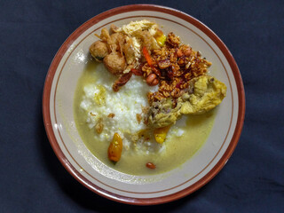 Bubur ayam. A warm Indonesian dish of chicken porridge with savory yellow sauce, chicken pieces, peanuts and orek tempeh served completely on a brown plate.
