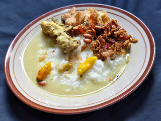 Bubur ayam. A warm Indonesian dish of chicken porridge with savory yellow sauce, chicken pieces, peanuts and orek tempeh served completely on a brown plate.