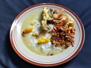 Bubur ayam. A warm Indonesian dish of chicken porridge with savory yellow sauce, chicken pieces, peanuts and orek tempeh served completely on a brown plate.