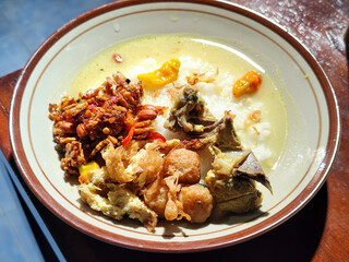 Bubur ayam. A warm Indonesian dish of chicken porridge with savory yellow sauce, chicken pieces, peanuts and orek tempeh served completely on a brown plate.