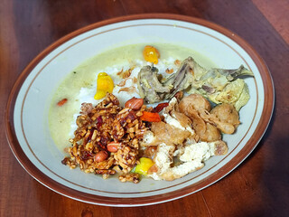 Bubur ayam. A warm Indonesian dish of chicken porridge with savory yellow sauce, chicken pieces, peanuts and orek tempeh served completely on a brown plate.