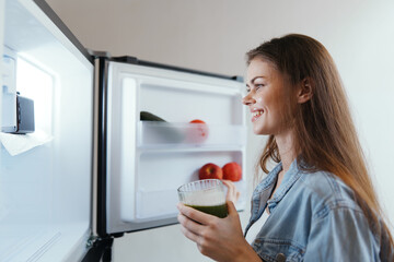 A young woman smiling while holding a green juice in front of her open refrigerator filled with...