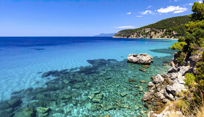 Obraz premium Clear turquoise ocean water with rocky coastline and green trees under blue sky